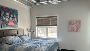 A bedroom with a bed, a window with blinds, and two artworks on the walls. - Brooklynn Suite - Pet Friendly - Dukes Place (Brandon)