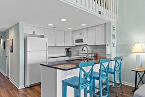 A modern kitchen with white cabinets, a central island with blue chairs, a microwave, and a coffee maker.