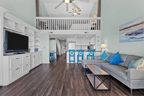 A spacious living area with a large flat-screen TV, a grey sofa with blue cushions, a wooden coffee table, and a dining area with blue chairs.