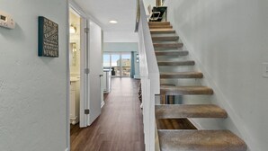 A staircase with a handrail leading to an upper floor. A door on the left leads to a hallway with a view of the outside. - OceanScape- Beach and Sun in Surf City! (Surf City)