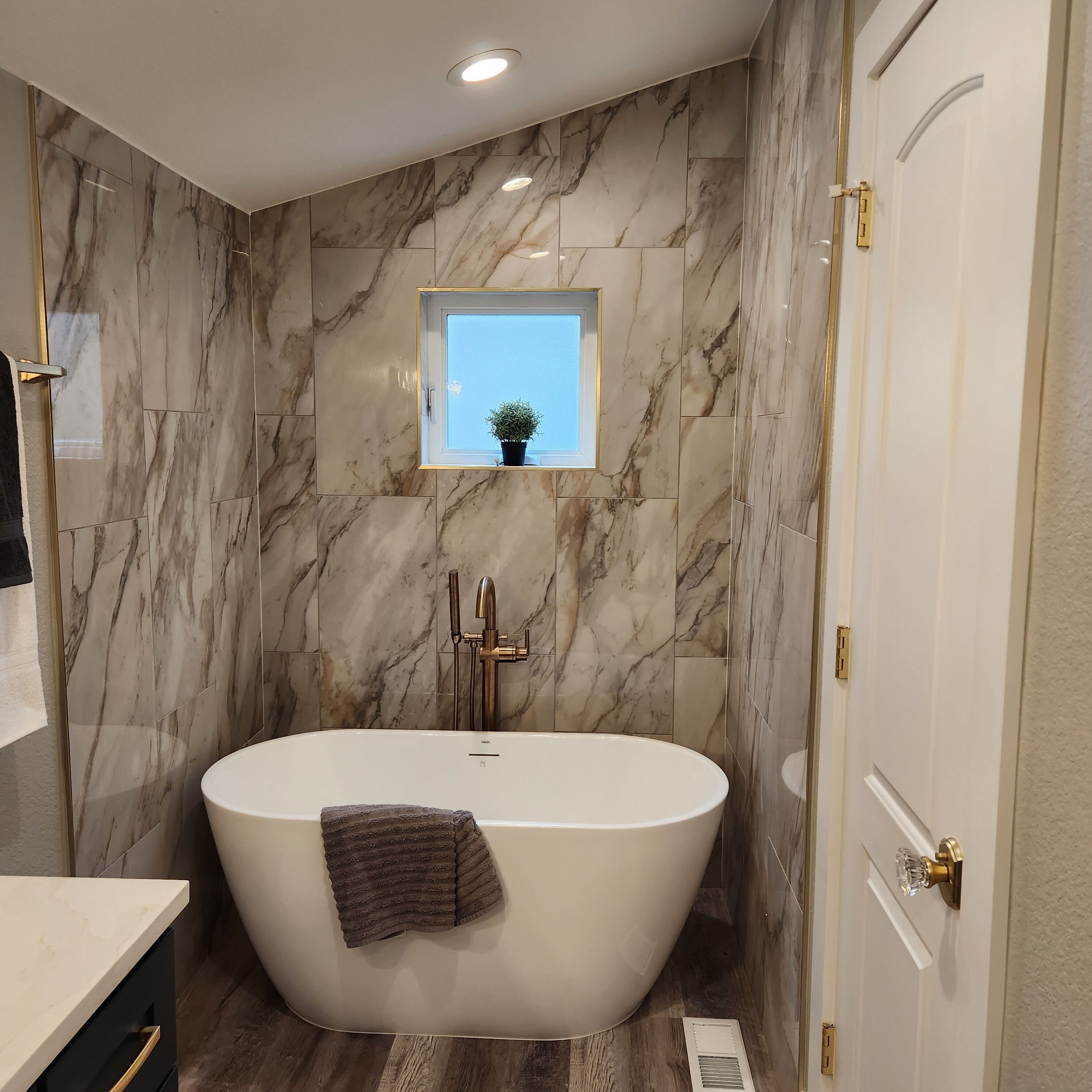 Een moderne badkamer met een vrijstaand bad, marmeren wanden, een klein raam en een deur met een gouden handvat.