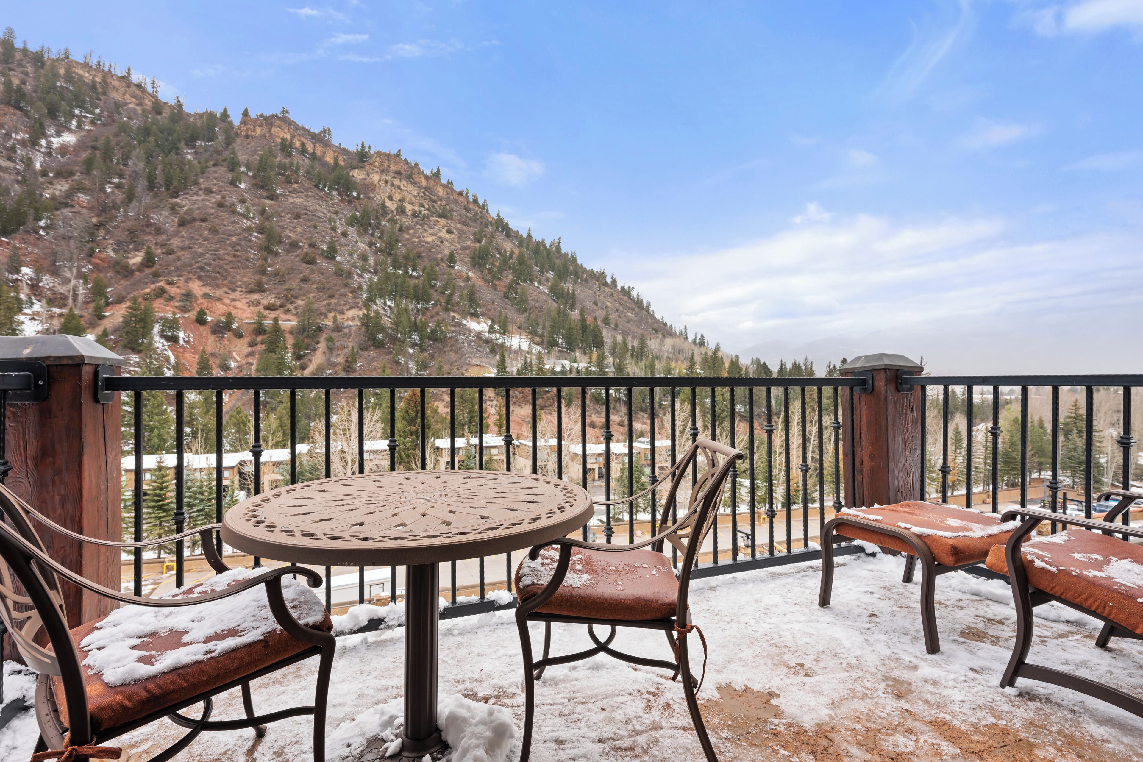A balcony with a round table and four chairs, snow on the ground, and a view of a mountainous landscape.