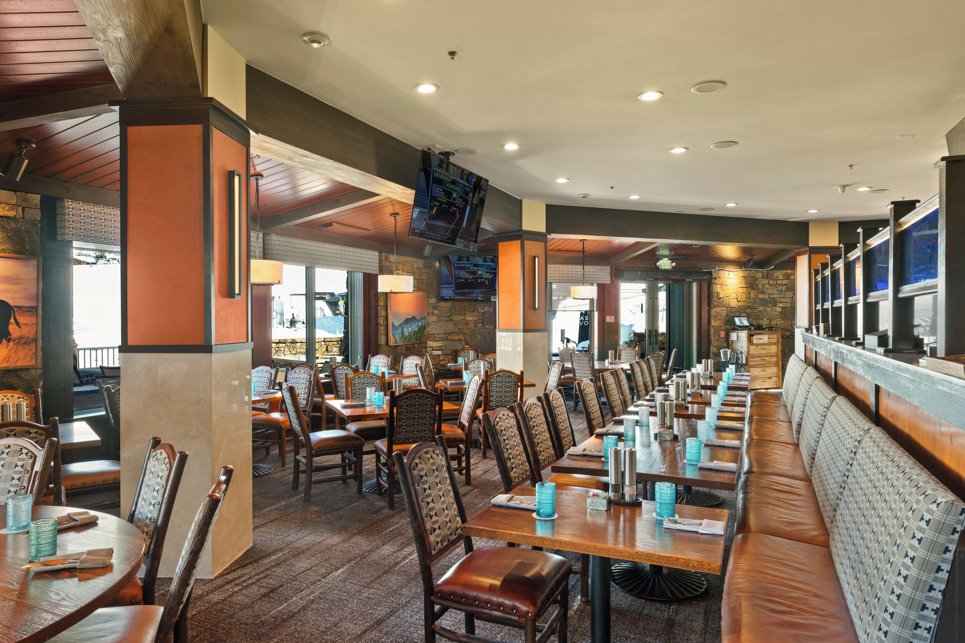 Ein Restaurant mit langen Gemeinschaftstischen und Sitzecken, Holzsesseln und einer Wand aus Naturstein.
