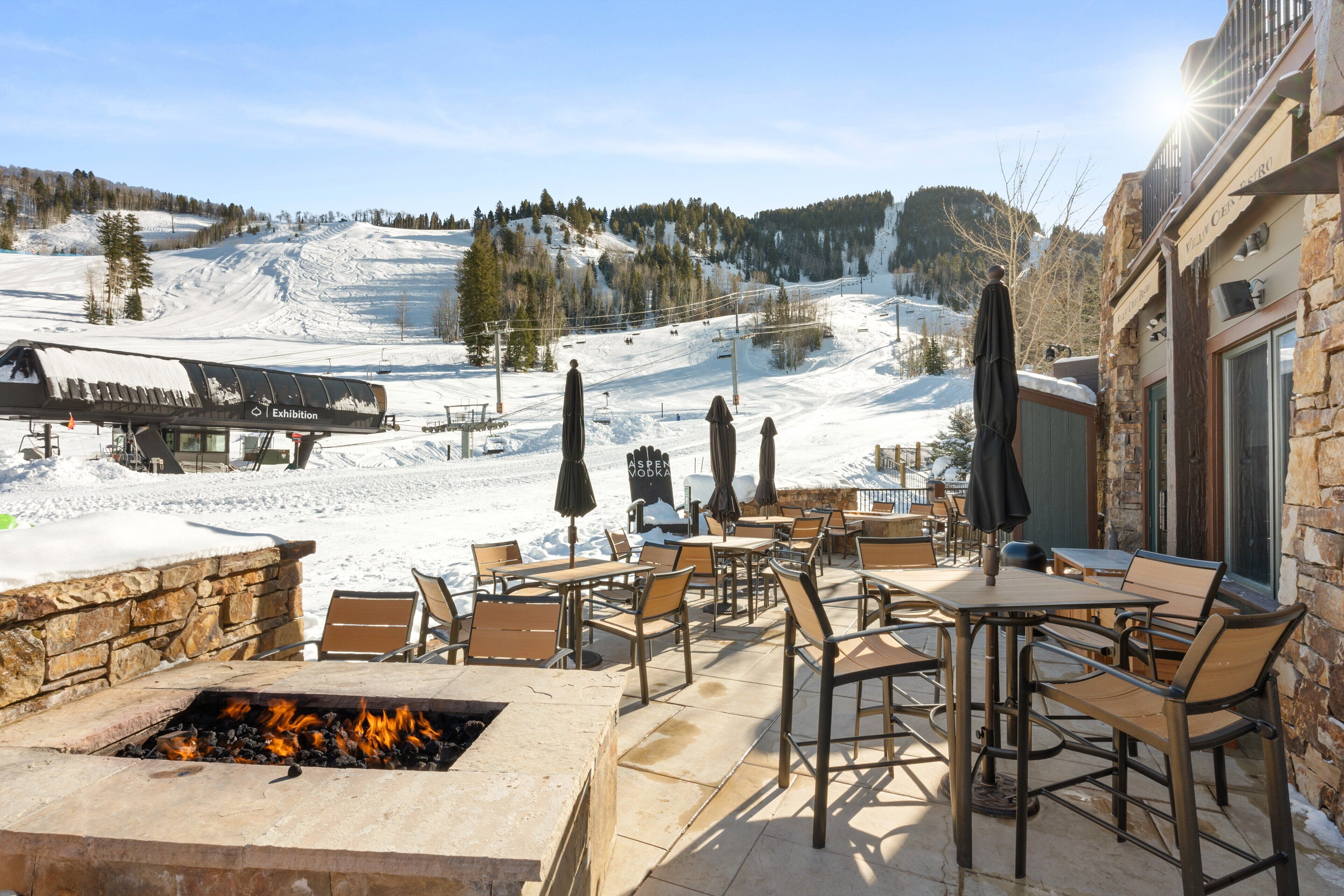 Un patio exterior con asientos de piedra y un pozo de fuego, con vistas a una zona nevada de montaña con un telesilla y un edificio identificado como "Hotel Centro".