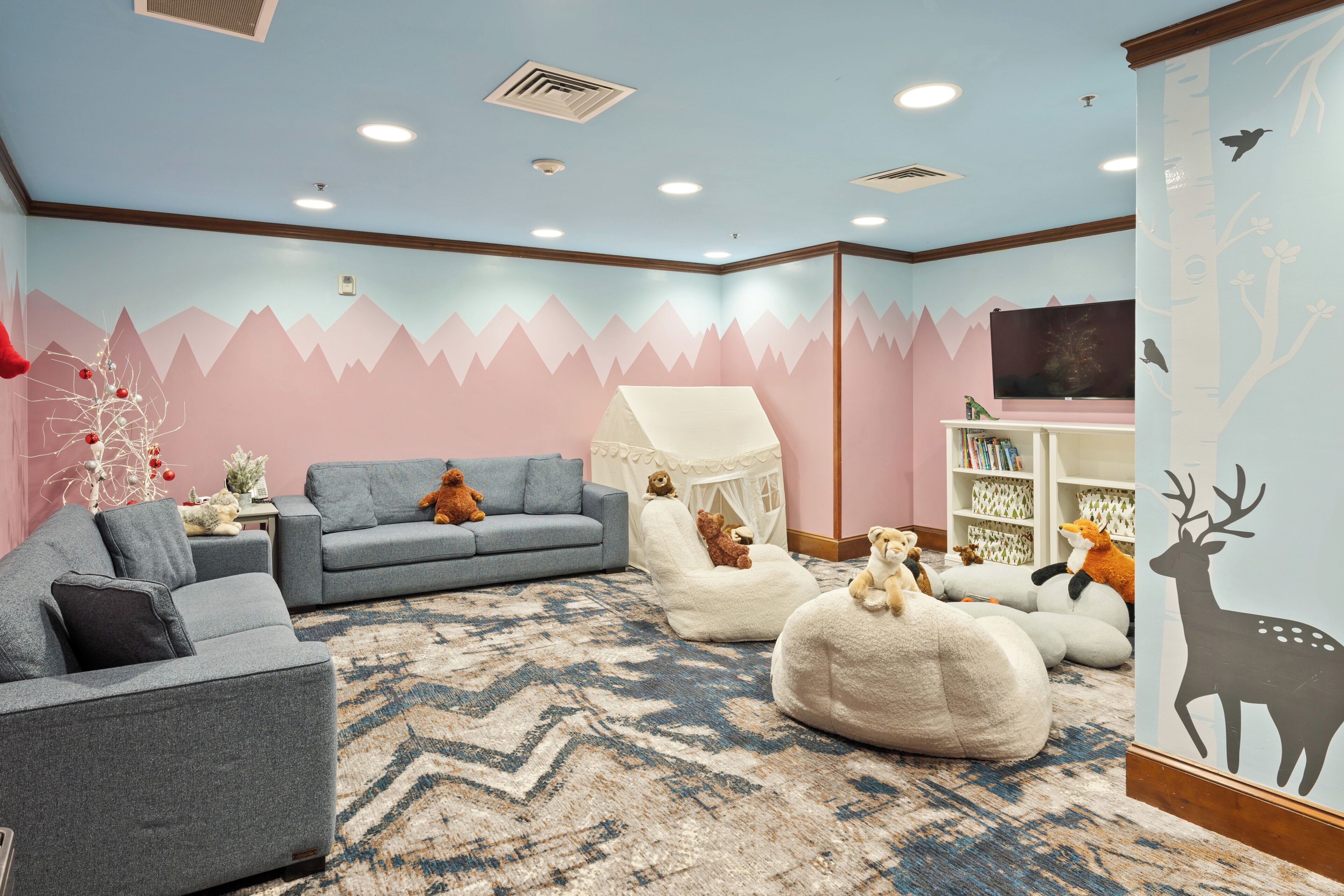 Una habitació acollidora amb un mural de muntanya blau i rosa, un sofà gris, una cadira blanca i una prestatgeria.