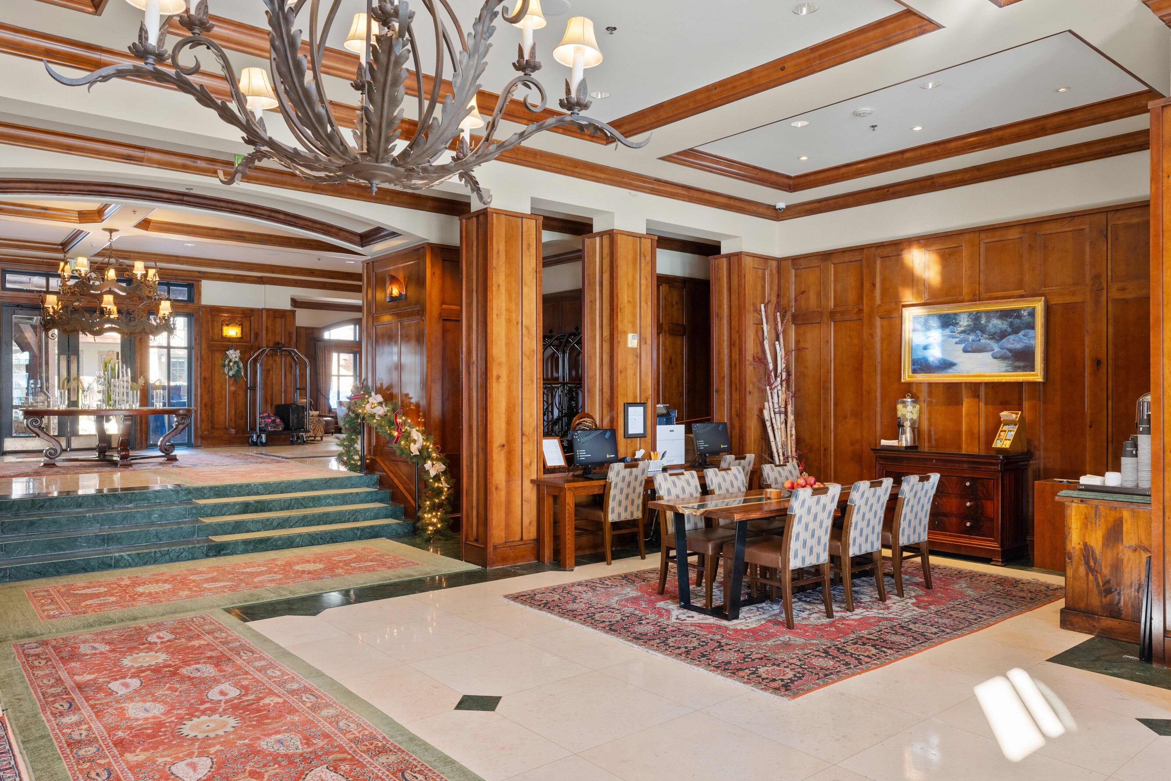 Un lobby espacioso con una gran araña de cristal, paneles de madera, un mostrador de recepción y una zona de comedor con sillas y una mesa.