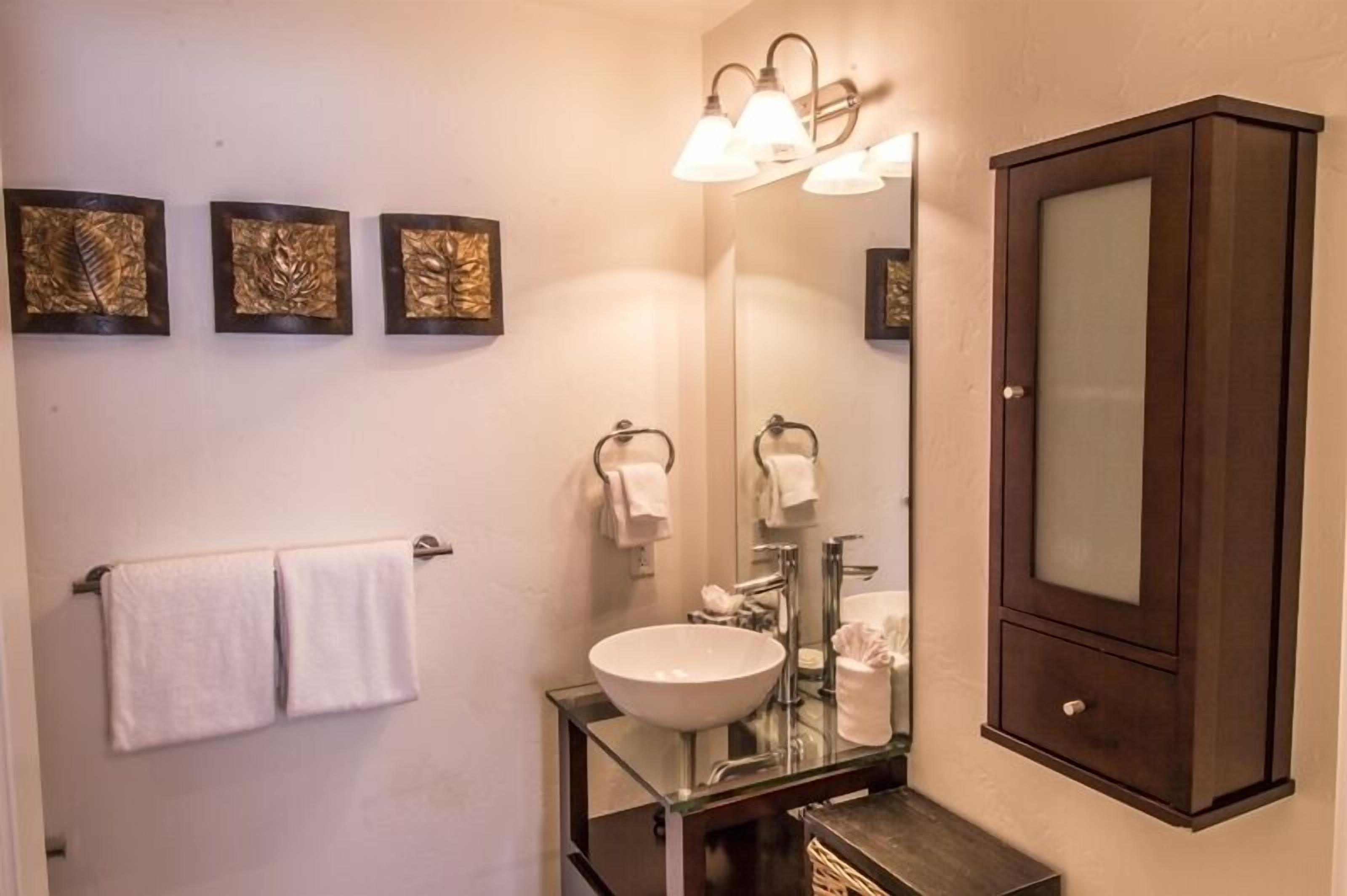 A bathroom with a glass sink, a large mirror, and three wall-mounted lights. There are three framed artworks above the sink and a towel rack with two towels.