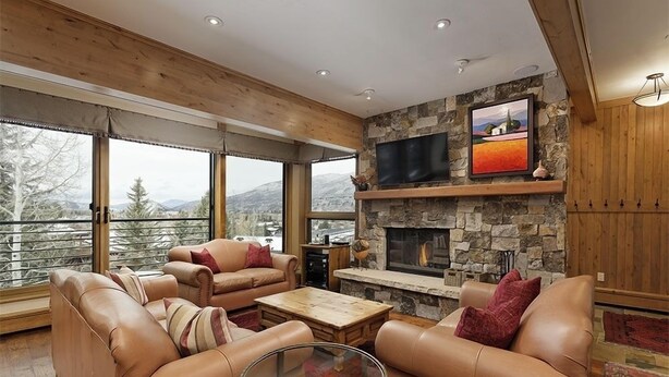 Ein gemütliches Wohnzimmer mit Ledersofas, einem Kamin aus Naturstein, einem Flachbildfernseher und großen Fenstern mit Blick auf die Berge.