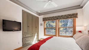 Ein Schlafzimmer mit einem Bett, einem an der Wand montierten Fernseher, einem Kleiderschrank und einem Fenster mit Blick auf Bäume.