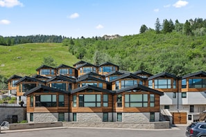 A modern multi-story building with large windows and a stone base, set against a hillside with a forested background.