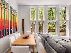 Un coin repas avec une table en bois et des chaises blanches, de grandes fenêtres donnant sur un paysage verdoyant, et des tableaux abstraits aux couleurs vives accrochés au mur.