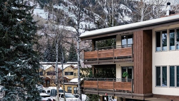 A modern building with a wooden balcony, surrounded by snow-covered trees and a mountainous backdrop.