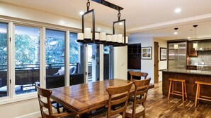 A modern kitchen with a wooden dining table and chairs, a bar area, and large windows offering a view of the outdoors.
