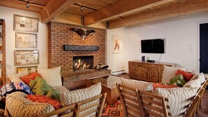 A cozy living room with a brick fireplace, wooden furniture, and a flat-screen TV mounted on the wall. - Cottonwoods Unit 1C | Downtown Condo with Upgraded Kitchen & Bathrooms (Aspen)