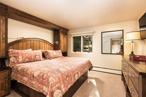 Ein Schlafzimmer mit Holz-Kopfteil, einem großen Bett mit floralem Bettzeug, einem Fenster mit Blick, einem Spiegel und einer Kommode mit Schubladen.
