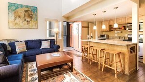 Ein modernes Wohnzimmer mit einem blauen Sofa, einem Holztisch und einem gemusterten Teppich. Im Hintergrund ist eine Küche mit Holzbarhocker und Pendelleuchten zu sehen.