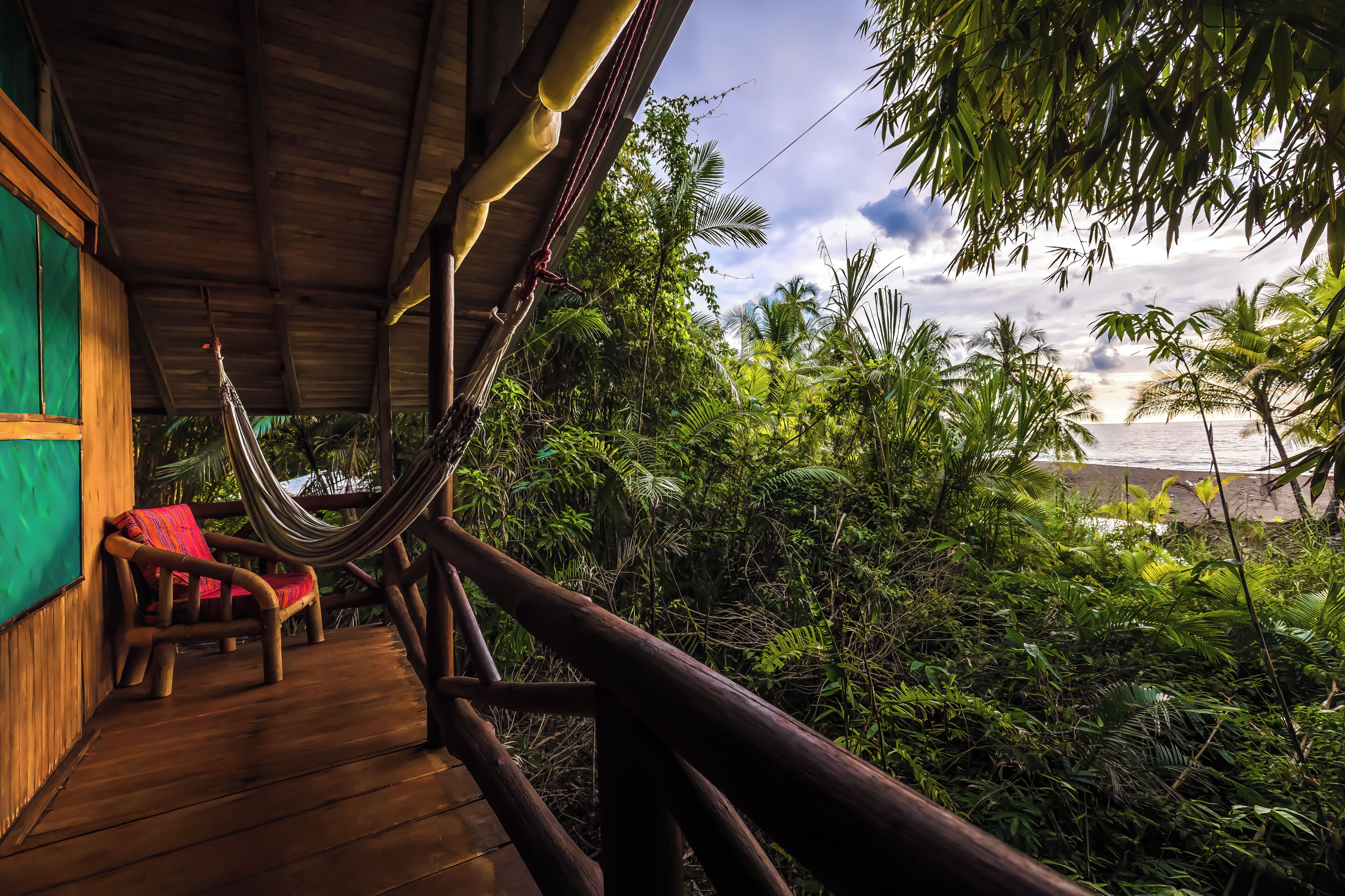 A wooden deck with a hammock and a chair, overlooking a lush tropical landscape with dense foliage and a view of the ocean.