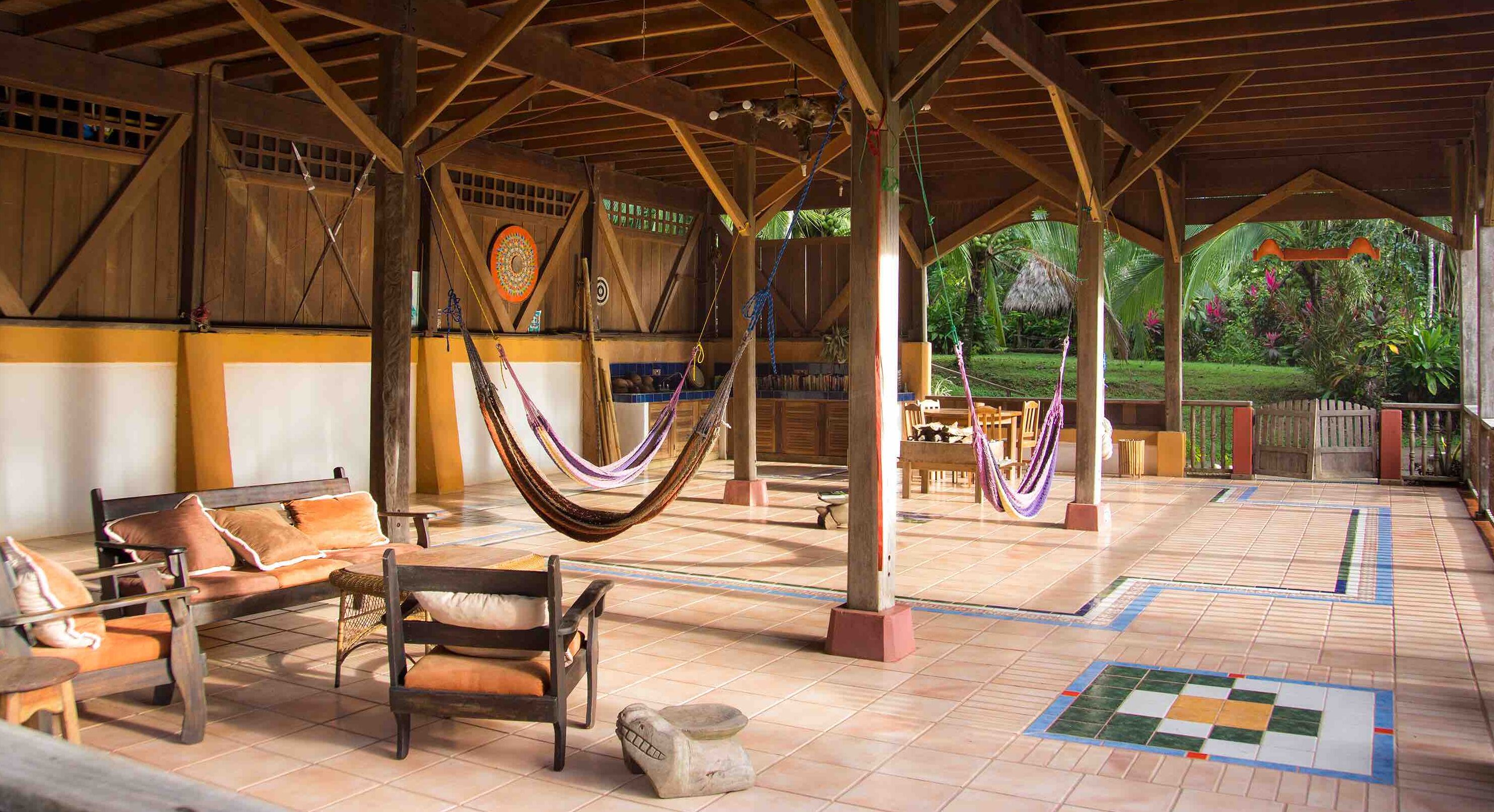 Un espacio al aire libre techado con mobiliario de madera, hamacas y suelo de baldosas.