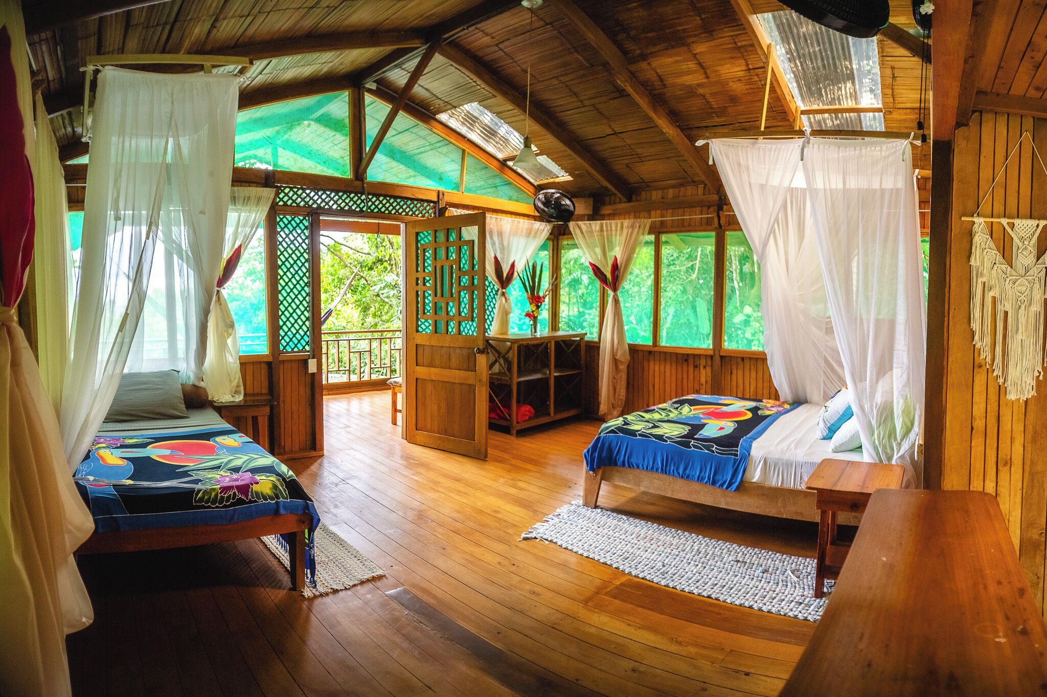 A wooden room with two beds, a wooden table, and a balcony with a view of greenery.
