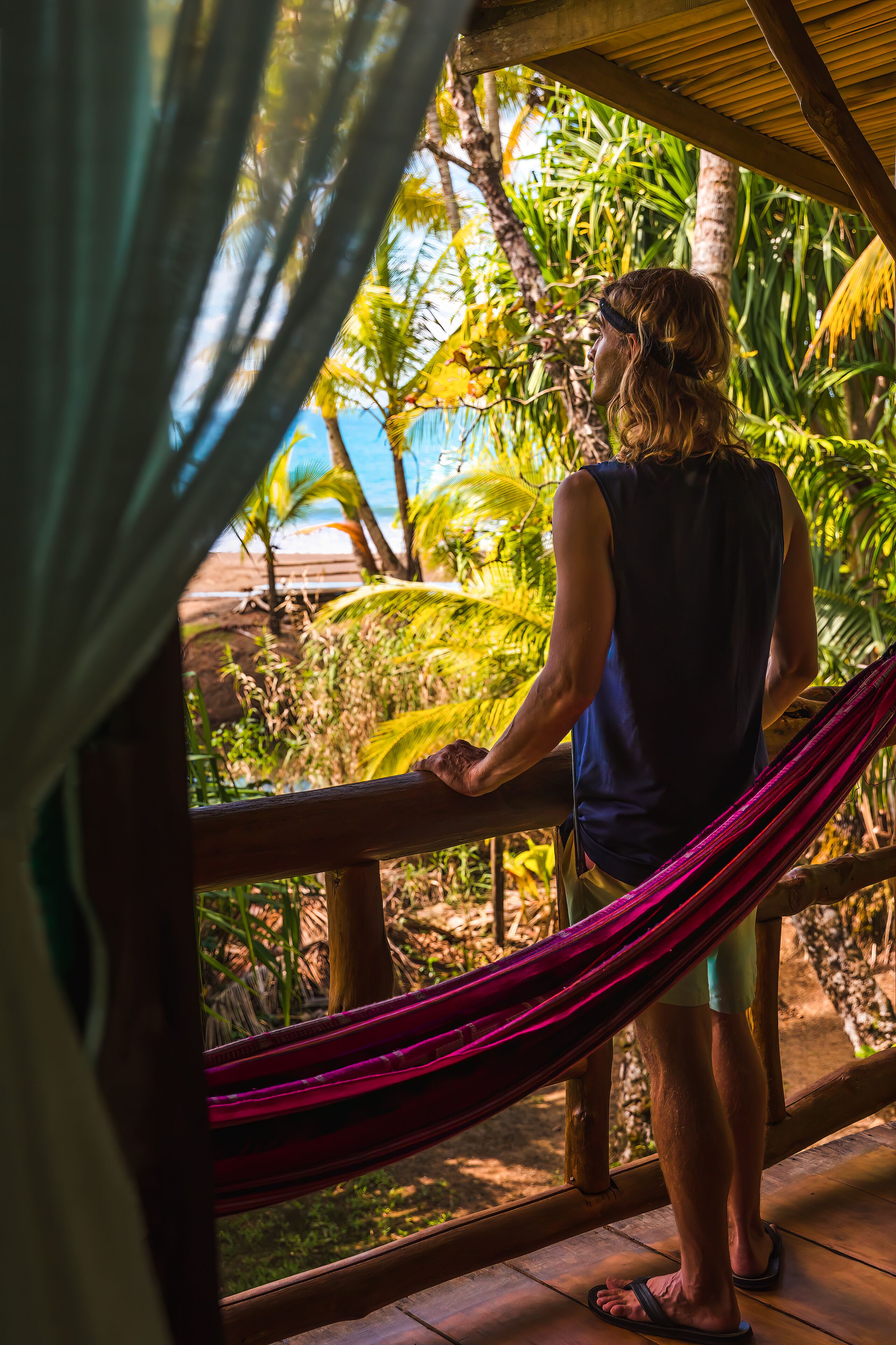 A person standing on a wooden deck, holding a hammock, with a view of a beach and palm trees.