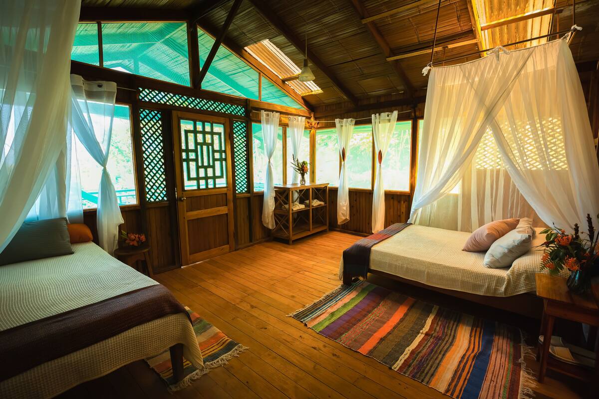 A room with two beds, wooden flooring, and a large window with a view of greenery.