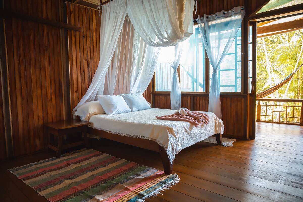 A wooden room with a bed, a nightstand, a colorful rug, and a large window with a view of trees.