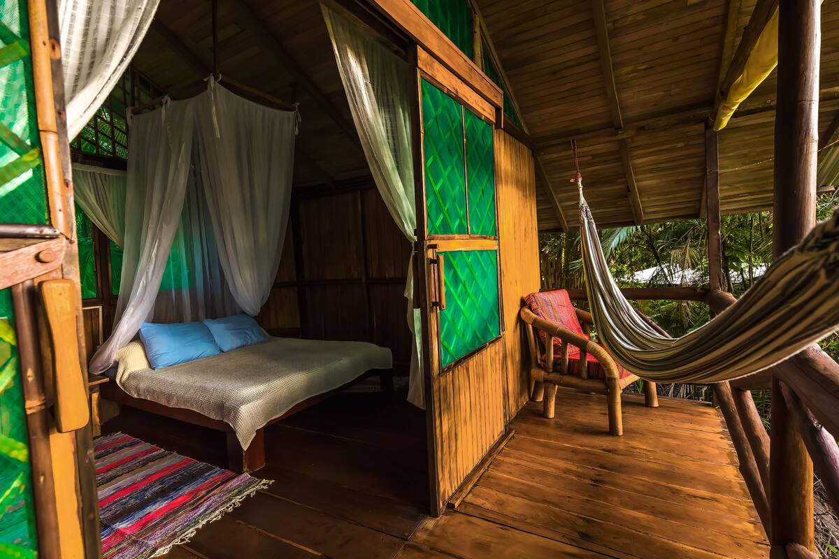 A wooden room with a bed, a hammock, and a view of greenery outside.
