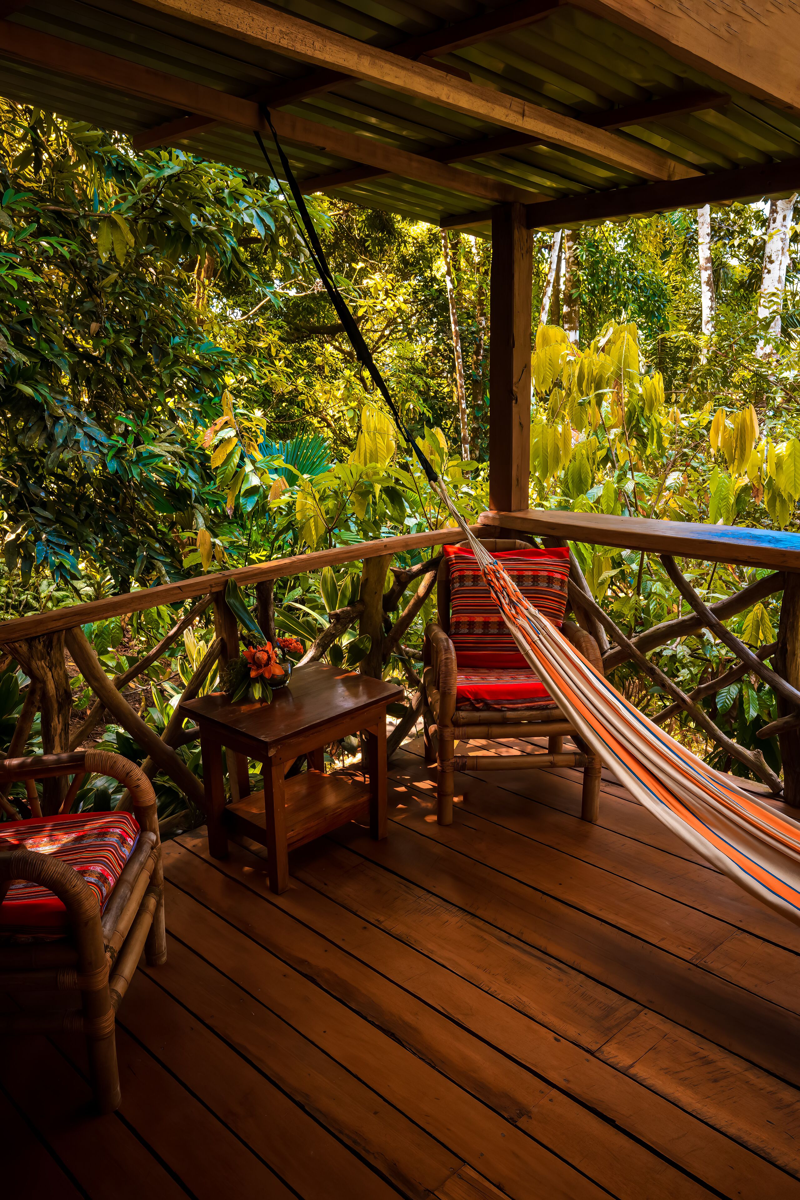 Une terrasse en bois avec un hamac, un fauteuil rouge rembourré et une petite table sur laquelle se trouve un panier de fruits.