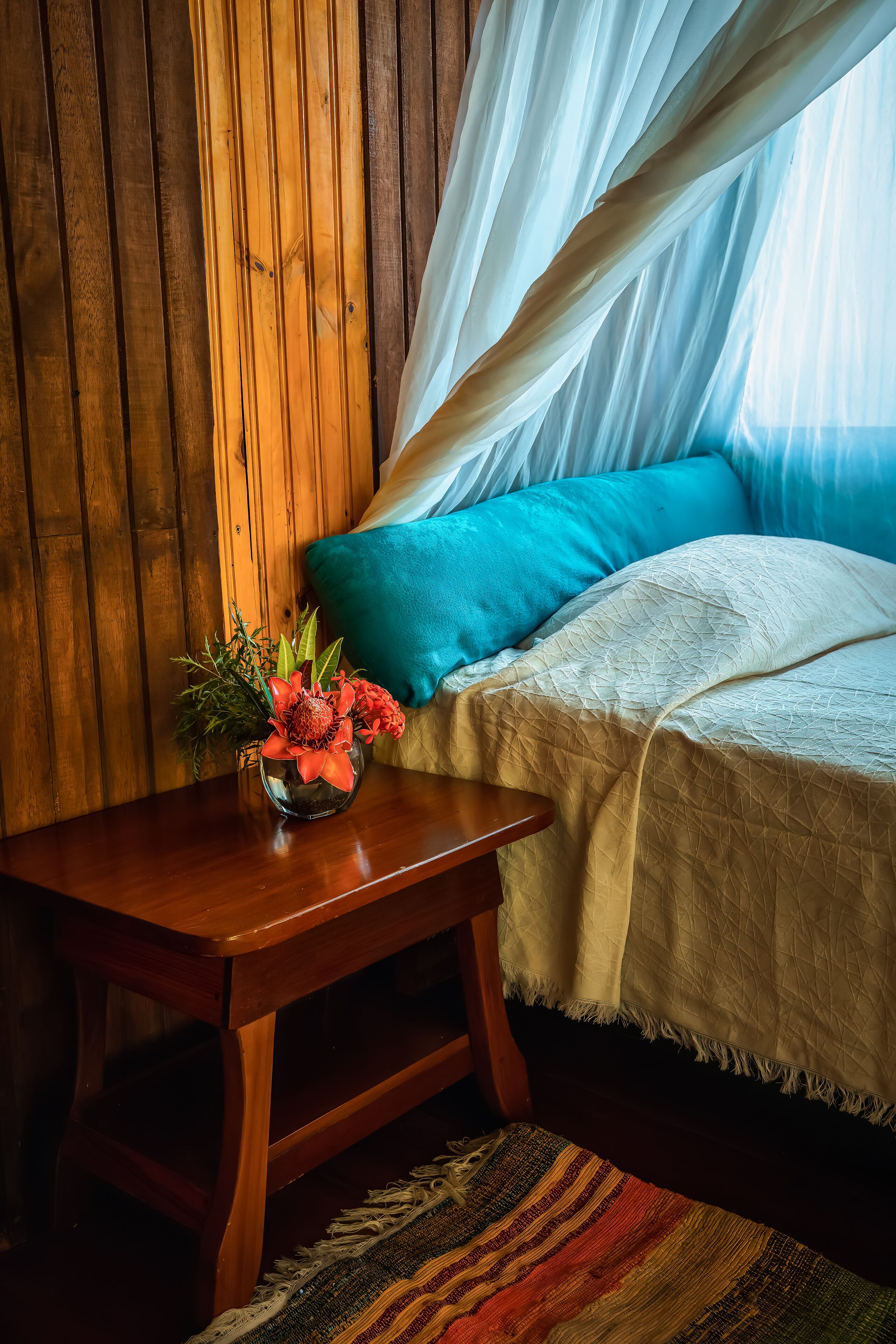 Una pared de madera, una cama con colcha blanca y almohadas azules, una mesita de noche de madera con un jarrón de flores y una alfombra tejida y colorida.