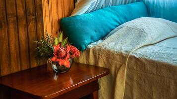 Una pared de madera, una cama con colcha blanca y almohadas azules, una mesita de noche de madera con un jarrón de flores y una alfombra tejida y colorida.