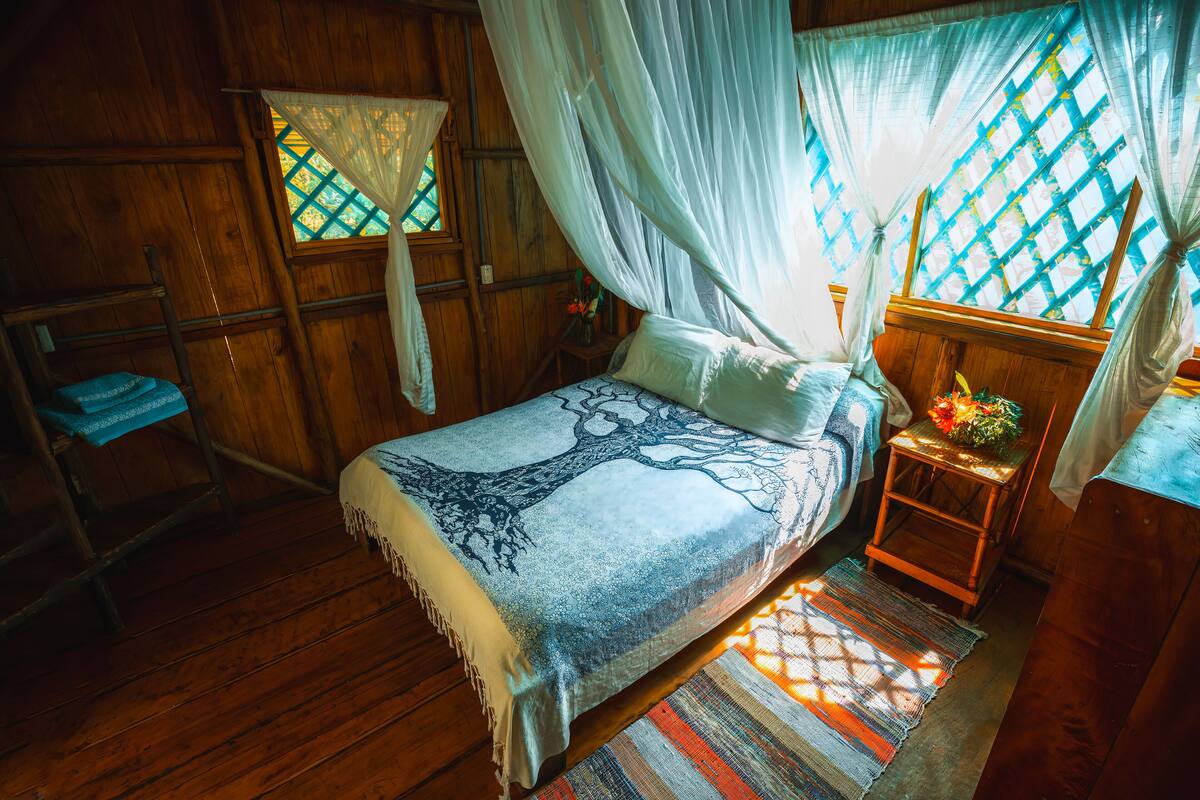 A wooden room with a bed, a small table with a vase, and a window with a lattice design.