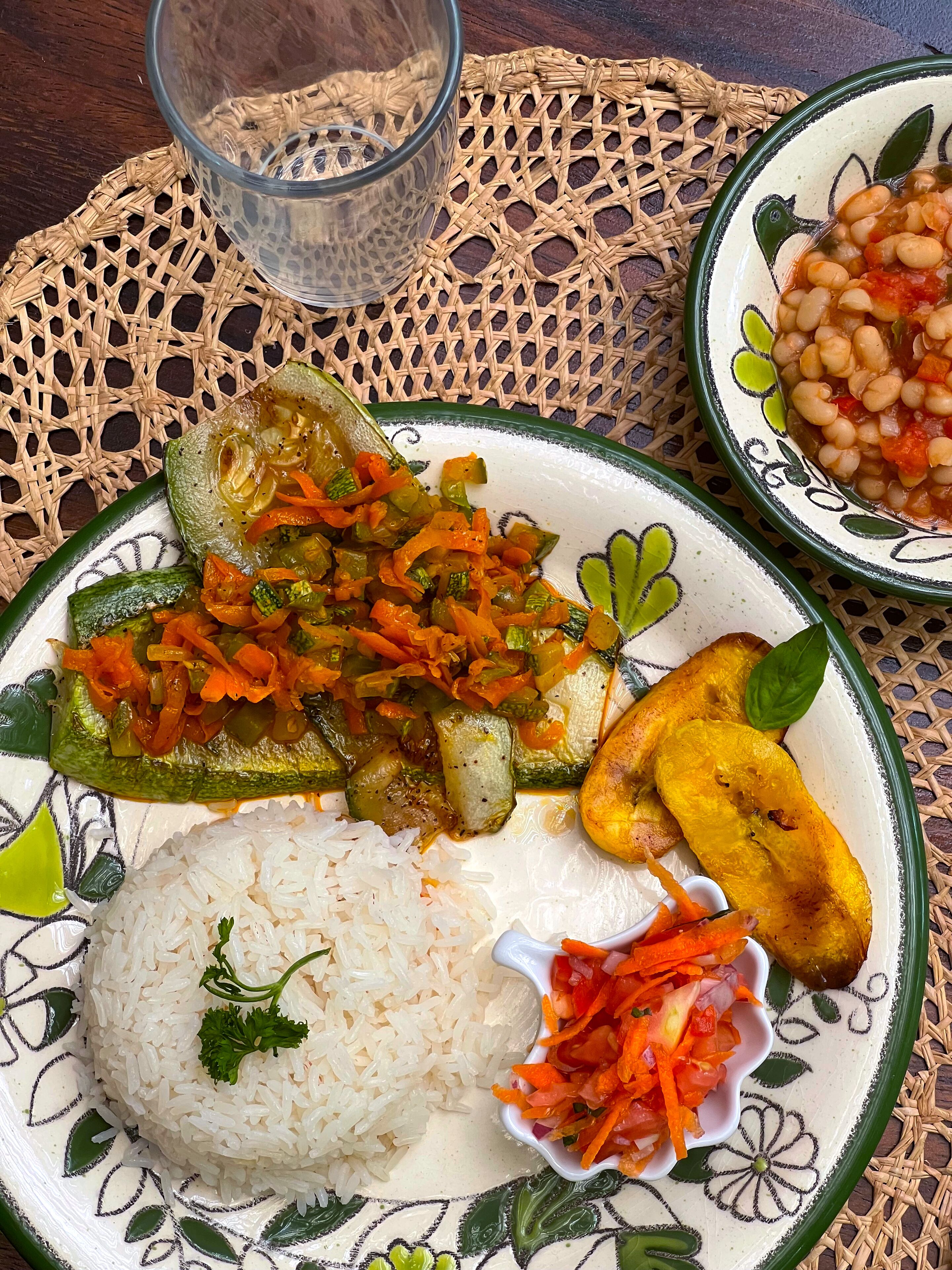 A plate with rice, a stuffed vegetable, a side of beans, and a glass of water on a woven placemat.