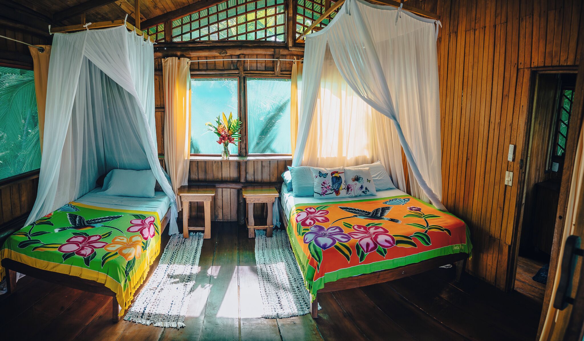 A room with two beds, each with a canopy, a small table, and a window with a view of greenery.