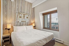 A bedroom with a large bed, wooden headboard, two bedside lamps, and a window with a view of a building. - Chateau Chaumont Unit 19 | Spacious Top-Floor Condo with Great Location (Aspen)