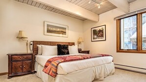 A bedroom with a large bed, two bedside tables, a nightstand, a wooden headboard, and two wall-mounted lamps.