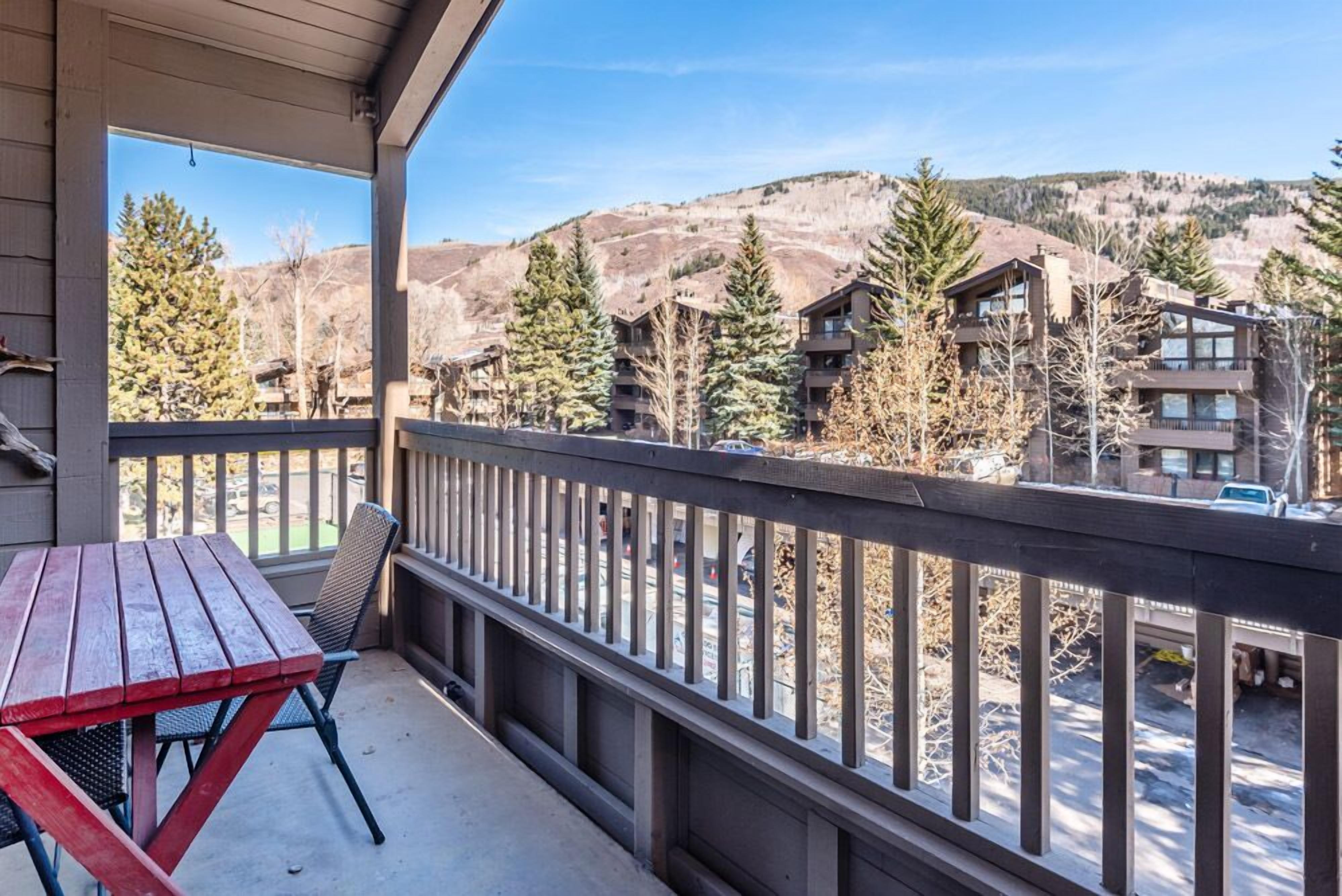 Un balcon doté d’une table et de chaises en bois, donnant sur un quartier résidentiel avec des maisons et des arbres.