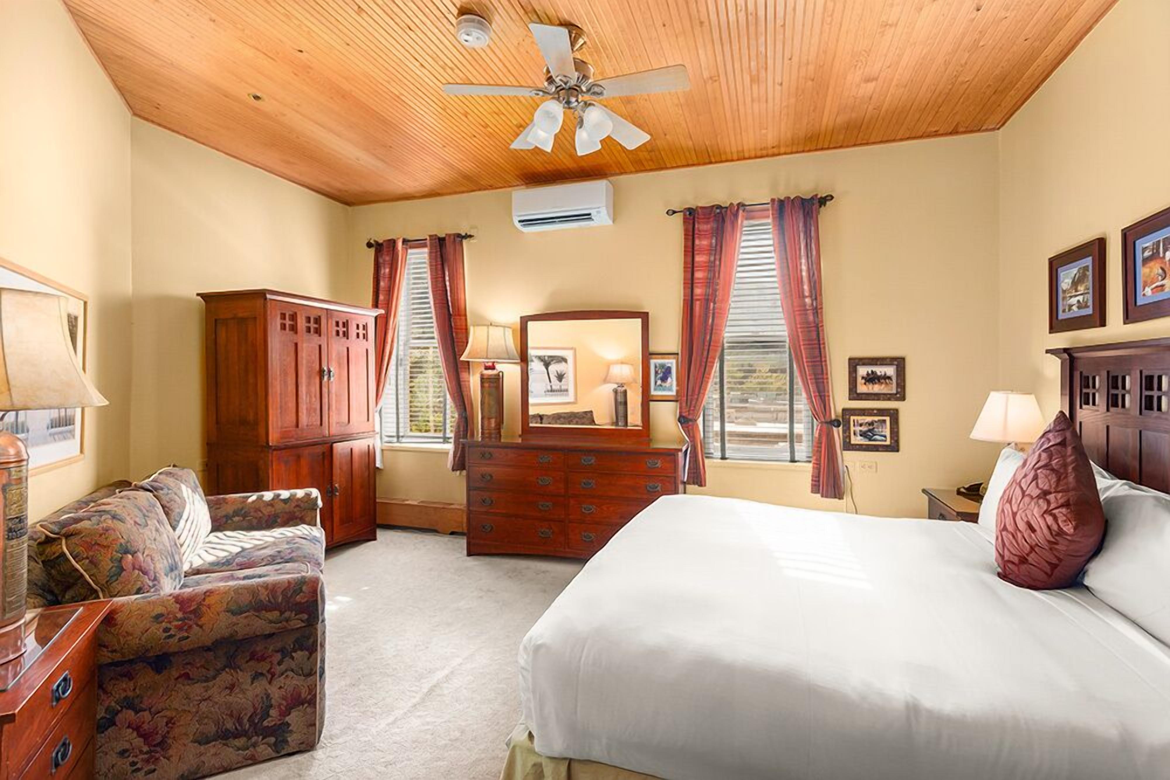 A bedroom with a bed, a wooden ceiling, a ceiling fan, a dresser, a mirror, and curtains.