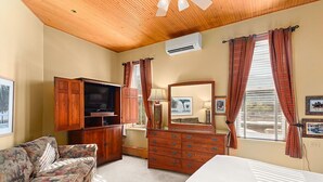 Una habitación con techo de madera, una cama grande, un aparador de madera, un televisor, un sofá y una ventana con cortinas.
