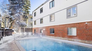 A swimming pool with a clear blue water surface, surrounded by a brick building with multiple windows and a covered patio area with chairs and tables.