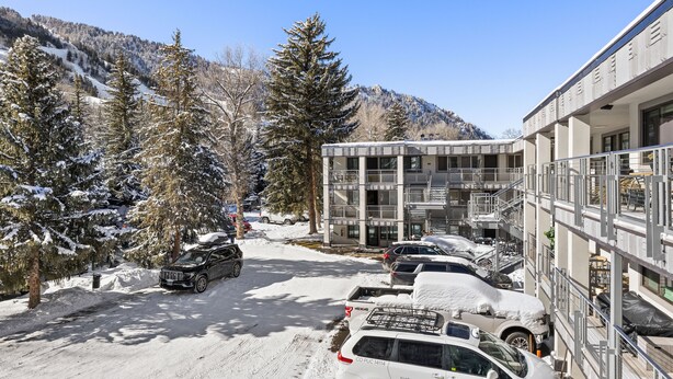 Un estacionamiento con coches cubiertos de nieve, un edificio de varios pisos y árboles nevados.