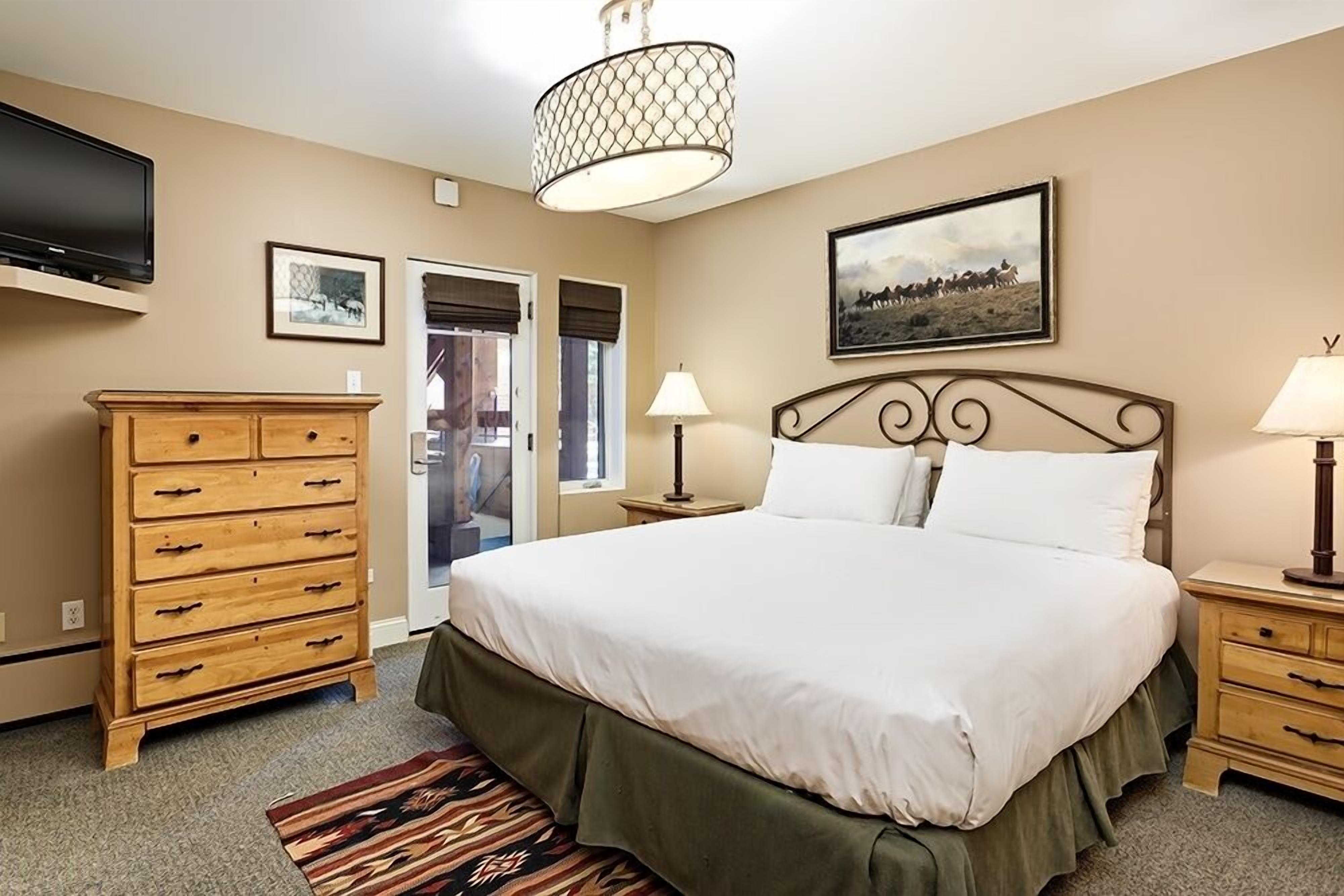 Een slaapkamer met een bed, een houten dressoir, twee nachtkastjes met lampen, een televisie aan de muur en een ingelijst schilderij boven het bed.