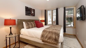 Ein Schlafzimmer mit einem hölzernen Bett, zwei Nachttischlampen, einem großen Fenster mit Jalousien und einem Wand-Fernseher.