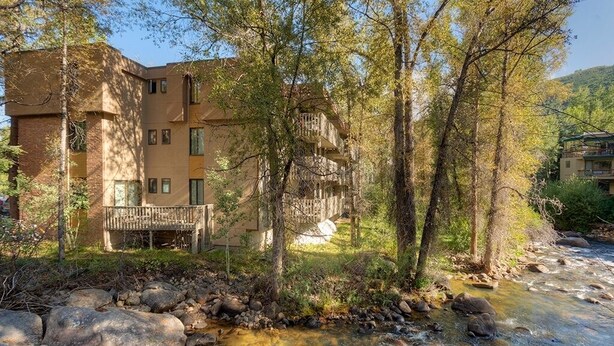A multi-story building with balconies, a stream in front, and trees. - Riverview Unit 8 | All the Essentials Riverside Condo with Access to Pool & Hot Tub (Aspen)