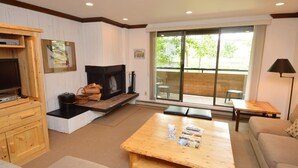 A cozy living room with a fireplace, wooden furniture, and a large window offering a view of the outdoors. - Riverview Unit 8 | All the Essentials Riverside Condo with Access to Pool & Hot Tub (Aspen)