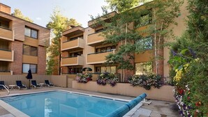 A swimming pool with lounge chairs, surrounded by a brick building with balconies and flower boxes. - Riverview Unit 8 | All the Essentials Riverside Condo with Access to Pool & Hot Tub (Aspen)
