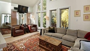 A spacious living room with a large sofa, a coffee table, and a TV mounted on the wall. - Old Hundred Unit 102 | Townhouse with Treetop Views & Excellent Location, 3 Blocks to Ski Slopes (Aspen)
