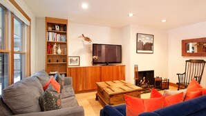 A living room with a grey sofa, a blue sofa, a wooden coffee table, a bookshelf, a television, a fireplace, and a large window.