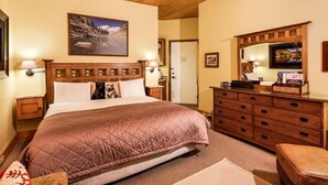 A bedroom with a large bed, wooden headboard, a painting of a mountain landscape, a dresser with drawers, and a television on a wooden cabinet. - Independence Square Unit 309 | Downtown Hotel Room with A/C in Aspen, Wet Bar & More (Aspen)