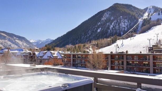 A hot tub on a balcony overlooking a mountainous area with buildings and a ski lift. - Independence Square 213 | Spacious Hotel Room with 2 Queen Beds, Wet Bar, and Sitting Area (Aspen)