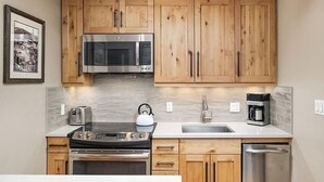 A kitchen with wooden cabinets, a stainless steel microwave, a stove, a kettle, a coffee maker, and a dishwasher. - Fasching Haus Unit 12 | Lower Level Deluxe Condo w/ Open Living Area & Gas Fireplace (Aspen)